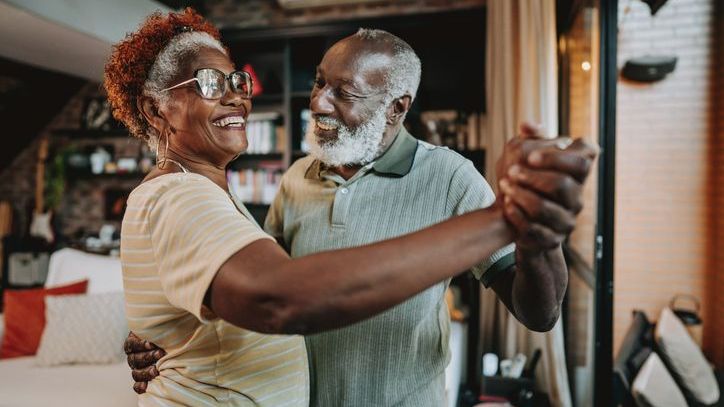 A happily retired couple that takes advantage of Social Security spousal benefits share a dance in their home.